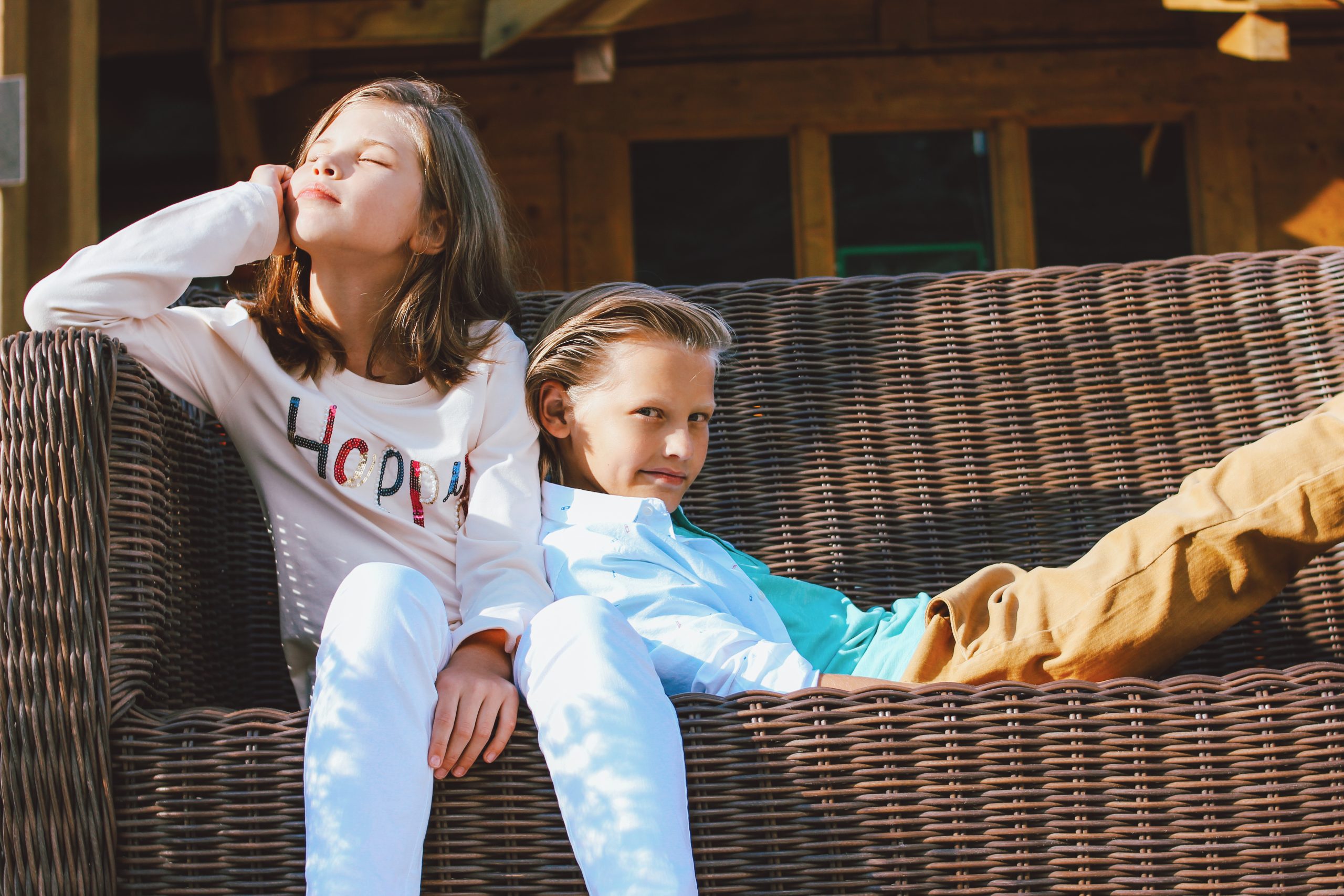 Attractive preteen children sister and brother, friends sitting on wicker sofa in backyard of the  wooden cottage